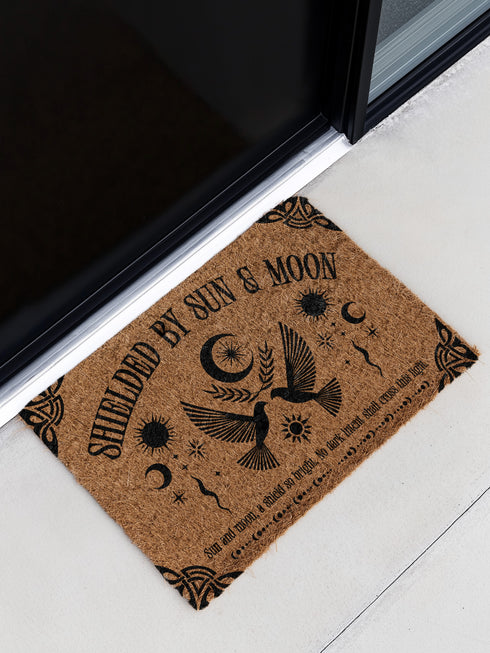 Natural coir doormat featuring two symmetrical doves beneath a crescent moon, surrounded by celestial and botanical accents. The phrase Shielded by Sun & Moon is stenciled above, with a protective incantation at the base. Non-slip vinyl backing for outdoor use.