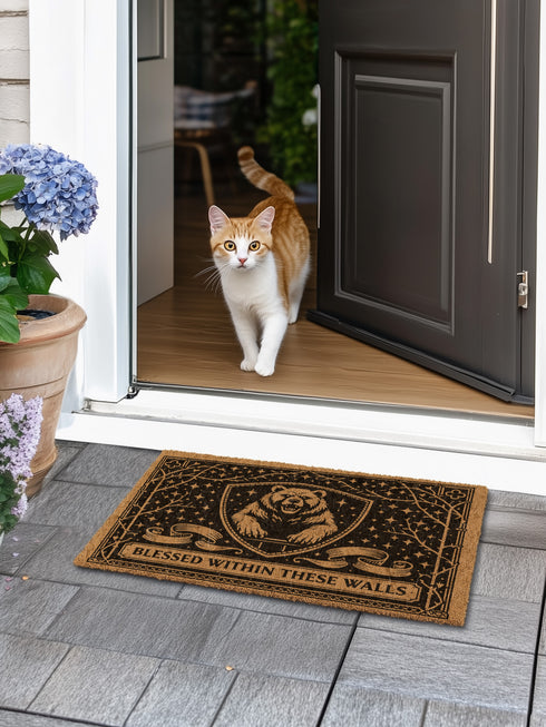 A rectangular eco-friendly coir doormat measuring 24 by 16 inches displayed on a stone porch. The natural golden-brown coconut fiber mat features a bold black graphic design of a heraldic bear head centered on a medieval knight's shield. Above and below the shield, Gothic-style typography reads BLESSED WITHIN THESE WALLS. The mat has a durable black vinyl non-slip backing. Ideal for Castlecore, Dark Academia, and Grimoire and Goblet style gothic home decor themes.