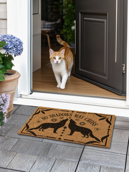Natural coir doormat featuring black silhouettes of two howling wolves in symmetrical formation, surrounded by celestial and botanical details. The phrase No Shadows May Cross is hand-stenciled above, with a non-slip vinyl backing for outdoor use.