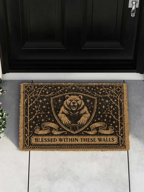 A rectangular eco-friendly coir doormat measuring 24 by 16 inches displayed on a stone porch. The natural golden-brown coconut fiber mat features a bold black graphic design of a heraldic bear head centered on a medieval knight's shield. Above and below the shield, Gothic-style typography reads BLESSED WITHIN THESE WALLS. The mat has a durable black vinyl non-slip backing. Ideal for Castlecore, Dark Academia, and Grimoire and Goblet style gothic home decor themes.