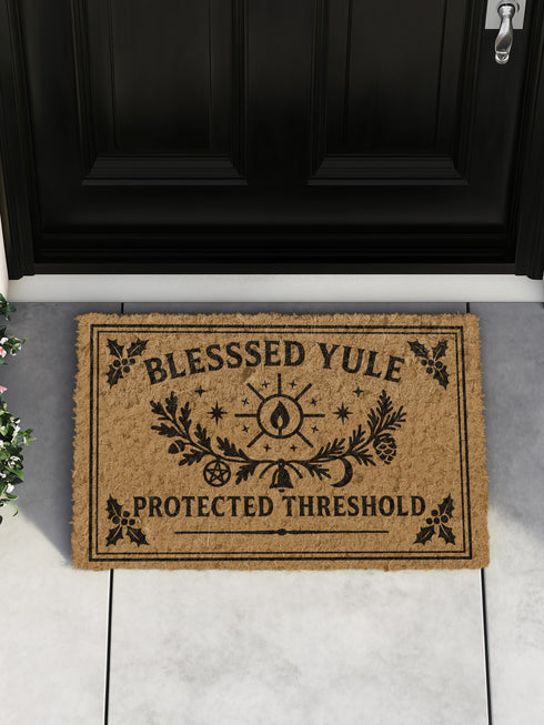 A rectangular tufted coir doormat with a non-slip black vinyl backing, features an intricate gothic botanical design in black. At the top, it reads "BLESSED YULE" with holly motifs in the corners. In the center, a laurel wreath, intertwined with a pentacle and a bell, encircles a radiant candle. Below the wreath rests a pinecone. At the bottom, the text "PROTECTED THRESHOLD" is flanked by holly. This pagan and witchy Grimoire and Goblet design is perfect for a gothic or dark academia entryway.