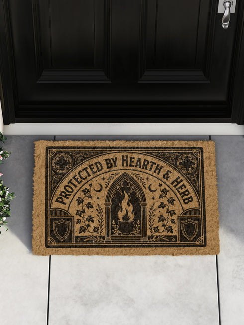 Rectangular coir doormat woven from natural brown coconut fiber with a bristly low pile. A bold black printed design fills the surface: a pointed archway frames a cast iron cauldron with rising flames at the center, flanked by winding ivy vines and small crescent moons. Arched lettering across the top reads PROTECTED BY HEARTH AND HERB. The four corners hold medieval shields and ornamental medallions, all enclosed by a thick black rectangular border with finished edges.