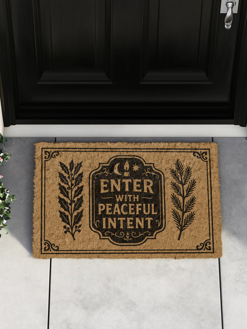 A rectangular tufted coir doormat from Grimoire and Goblet. The mat is a natural, textured brown, providing a high-contrast background for the black, gothic-style design. In the center, a decorative plaque, reminiscent of a vintage grimoire page, features the words "ENTER WITH PEACEFUL INTENT" in a serif font. Above the text is a small celestial crescent moon and star. Flanking the central plaque are two large, symmetrical botanical laurels or fern branches.