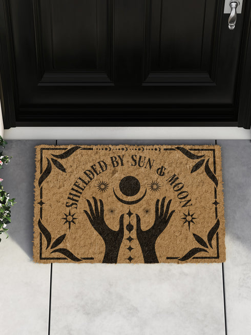 Natural coir doormat featuring black silhouettes of two open hands reaching toward a crescent moon and sun, surrounded by celestial and botanical details. The phrase Shielded by Sun & Moon is hand-stenciled above, with a non-slip vinyl backing for outdoor use.