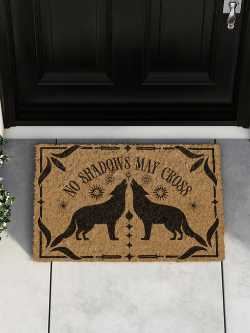 Natural coir doormat featuring black silhouettes of two howling wolves in symmetrical formation, surrounded by celestial and botanical details. The phrase No Shadows May Cross is hand-stenciled above, with a non-slip vinyl backing for outdoor use.