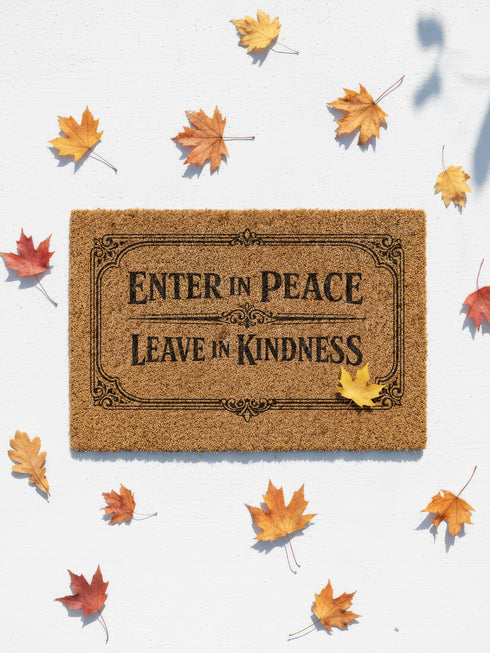 A high-angle close-up view of a Grimoire and Goblet luxury coir doormat. The mat features the phrase Enter in Peace Leave in Kindness painted in bold, Gothic-style black typography against the natural golden-brown texture of coconut husks. The text is enclosed in an intricate Victorian ornamental border with filigree corner flourishes. The texture appears coarse, durable, and dense, typical of high-quality tufted coconut fiber entry mats. The design evokes a Dark Academia and witchy aesthetic.