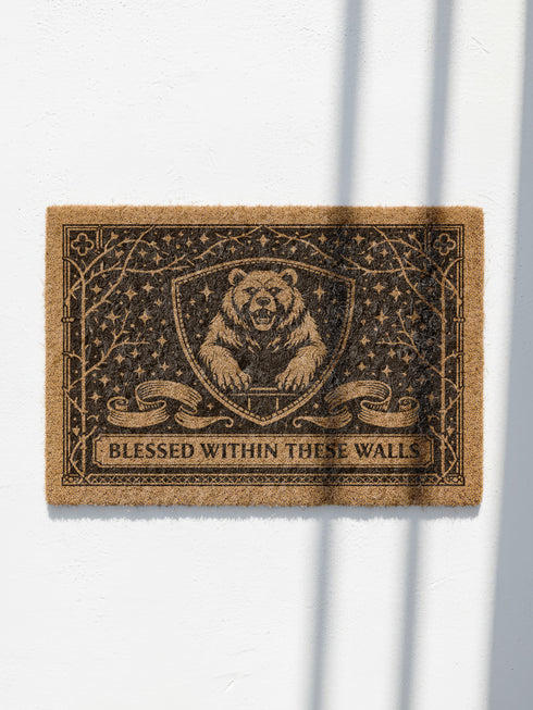A rectangular eco-friendly coir doormat measuring 24 by 16 inches displayed on a stone porch. The natural golden-brown coconut fiber mat features a bold black graphic design of a heraldic bear head centered on a medieval knight's shield. Above and below the shield, Gothic-style typography reads BLESSED WITHIN THESE WALLS. The mat has a durable black vinyl non-slip backing. Ideal for Castlecore, Dark Academia, and Grimoire and Goblet style gothic home decor themes.