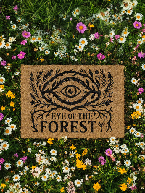 Rectangular coir doormat featuring black-ink artwork of a large all-seeing eye surrounded by branches, leaves, and forest flora. Beneath the design, the phrase “Eye of the Forest” is printed in bold serif font. Made from natural coconut coir with a vinyl backing, the mat measures 24 by 16 inches and is suitable for outdoor entryways.