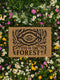 Rectangular coir doormat featuring black-ink artwork of a large all-seeing eye surrounded by branches, leaves, and forest flora. Beneath the design, the phrase “Eye of the Forest” is printed in bold serif font. Made from natural coconut coir with a vinyl backing, the mat measures 24 by 16 inches and is suitable for outdoor entryways.