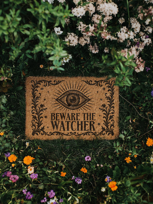 Coir doormat printed with “Beware the Watcher” in black ink, featuring a central all-seeing eye, ornate floral border, and star accents. Shown on outdoor stairs and entryways. Made from natural coconut fiber with black vinyl backing. A bold, mystical gift for witches, goth home lovers, and spiritual housewarmings.