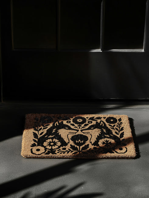 A rectangular, eco-friendly coir doormat with a natural, light brown tufted coconut fiber. Features a striking, symmetrical black folk-art design. In the center, two foxes face each other, noses touching, in a heraldic pose. They are surrounded by a dense, Scandinavian-style botanical border of stylized flowers, leaves, and branches, evoking a mystical woodland or dark cottagecore aesthetic. This witchy, Art Nouveau-inspired design serves as an elegant, gothic, or pagan piece of entryway decor.