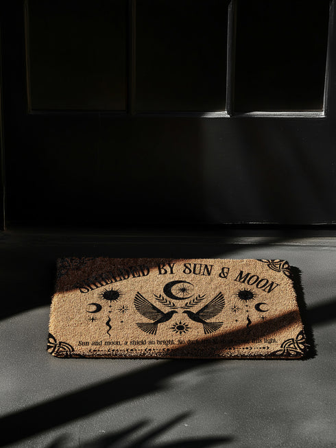 Natural coir doormat featuring two symmetrical doves beneath a crescent moon, surrounded by celestial and botanical accents. The phrase Shielded by Sun & Moon is stenciled above, with a protective incantation at the base. Non-slip vinyl backing for outdoor use.