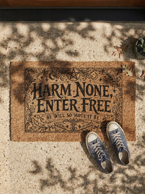 A rectangular natural coir doormat featuring the Wiccan Rede "An' It Harm None, Do What Ye Will" in a gothic dark academia style. The central typography is surrounded by symmetrical botanical flourishes and mystical symbols printed in high-contrast black ink. The 2D flat print mimics the look of a vintage grimoire page on the coarse, tan coconut fiber surface. The mat has a durable black vinyl backing and is pictured at a front door as a protective threshold guardian.