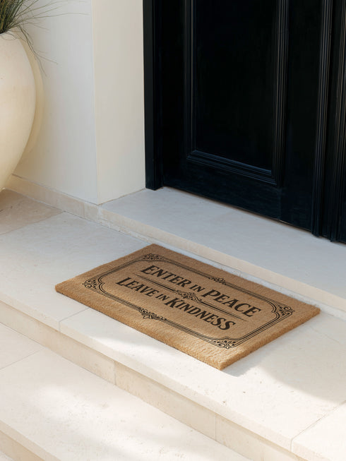 A high-angle close-up view of a Grimoire and Goblet luxury coir doormat. The mat features the phrase Enter in Peace Leave in Kindness painted in bold, Gothic-style black typography against the natural golden-brown texture of coconut husks. The text is enclosed in an intricate Victorian ornamental border with filigree corner flourishes. The texture appears coarse, durable, and dense, typical of high-quality tufted coconut fiber entry mats. The design evokes a Dark Academia and witchy aesthetic.