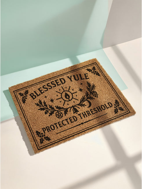 A rectangular tufted coir doormat with a non-slip black vinyl backing, features an intricate gothic botanical design in black. At the top, it reads "BLESSED YULE" with holly motifs in the corners. In the center, a laurel wreath, intertwined with a pentacle and a bell, encircles a radiant candle. Below the wreath rests a pinecone. At the bottom, the text "PROTECTED THRESHOLD" is flanked by holly. This pagan and witchy Grimoire and Goblet design is perfect for a gothic or dark academia entryway.