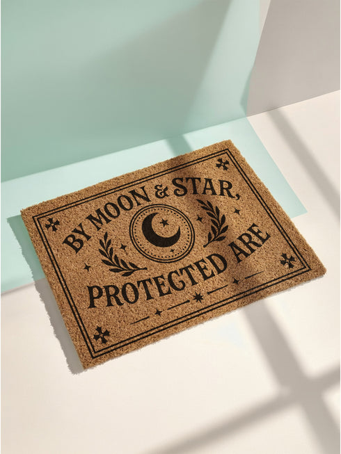 A rectangular, natural brown tufted coir doormat with a coarse, bristly texture designed to remove debris from shoes. Printed in bold black ink, the design features a central celestial emblem with a crescent moon and a single star, encircled by a decorative dotted border. Flanking this emblem are two symmetrical laurel leaf branches. The words "BY MOON & STAR," are in an arch above, with "PROTECTED ARE" in a straight line below, all in a serif, gothic-style font.