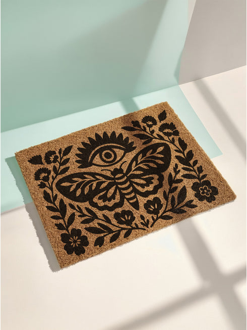 A rectangular, natural brown coir doormat with a coarse, tufted texture. The design is printed in solid black ink in a folk-art, block-print style. In the center is a large, stylized death's-head moth with intricate patterns on its wings. Above the moth is a central all-seeing eye with radiating lashes. The entire central image is enclosed by a symmetrical botanical frame of stylized flowers, stems, and leaves, creating a decorative border. The doormat has a non-slip black vinyl backing.