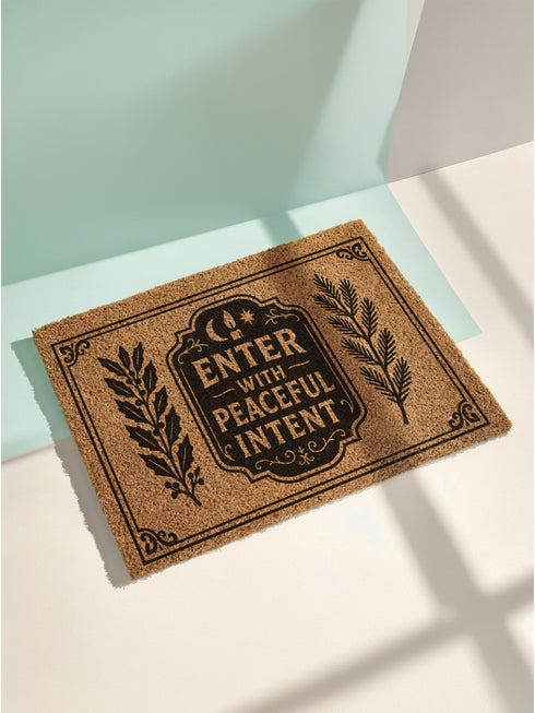 A rectangular tufted coir doormat from Grimoire and Goblet. The mat is a natural, textured brown, providing a high-contrast background for the black, gothic-style design. In the center, a decorative plaque, reminiscent of a vintage grimoire page, features the words "ENTER WITH PEACEFUL INTENT" in a serif font. Above the text is a small celestial crescent moon and star. Flanking the central plaque are two large, symmetrical botanical laurels or fern branches.