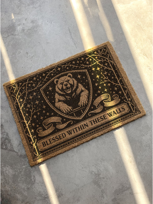 A rectangular eco-friendly coir doormat measuring 24 by 16 inches displayed on a stone porch. The natural golden-brown coconut fiber mat features a bold black graphic design of a heraldic bear head centered on a medieval knight's shield. Above and below the shield, Gothic-style typography reads BLESSED WITHIN THESE WALLS. The mat has a durable black vinyl non-slip backing. Ideal for Castlecore, Dark Academia, and Grimoire and Goblet style gothic home decor themes.