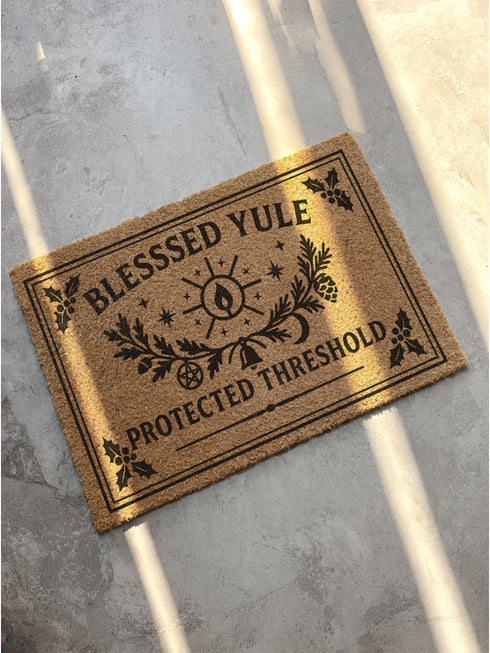 A rectangular tufted coir doormat with a non-slip black vinyl backing, features an intricate gothic botanical design in black. At the top, it reads "BLESSED YULE" with holly motifs in the corners. In the center, a laurel wreath, intertwined with a pentacle and a bell, encircles a radiant candle. Below the wreath rests a pinecone. At the bottom, the text "PROTECTED THRESHOLD" is flanked by holly. This pagan and witchy Grimoire and Goblet design is perfect for a gothic or dark academia entryway.