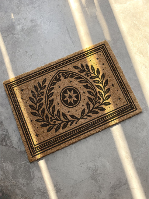 A rectangular natural coir doormat with a durable, non-slip black vinyl backing. The surface features an intricate black gothic-style design centered around an eight-pointed celestial star. This mystical symbol is encircled by a pointed arch formed from two botanical laurel branches, symbolizing wisdom and protection. The central motif is framed by a double border with geometric details and smaller stars. A perfect piece of spiritual home decor for a pagan or mystical entryway.