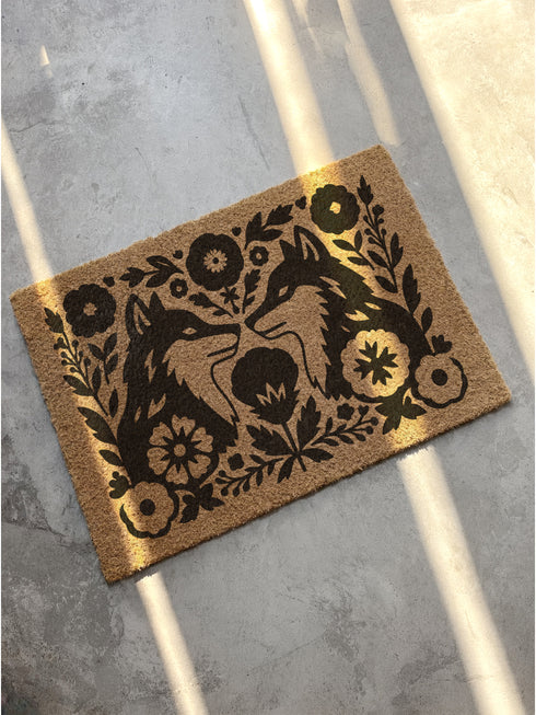A rectangular, eco-friendly coir doormat with a natural, light brown tufted coconut fiber. Features a striking, symmetrical black folk-art design. In the center, two foxes face each other, noses touching, in a heraldic pose. They are surrounded by a dense, Scandinavian-style botanical border of stylized flowers, leaves, and branches, evoking a mystical woodland or dark cottagecore aesthetic. This witchy, Art Nouveau-inspired design serves as an elegant, gothic, or pagan piece of entryway decor.