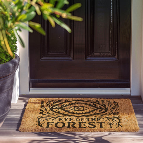 Rectangular coir doormat featuring black-ink artwork of a large all-seeing eye surrounded by branches, leaves, and forest flora. Beneath the design, the phrase “Eye of the Forest” is printed in bold serif font. Made from natural coconut coir with a vinyl backing, the mat measures 24 by 16 inches and is suitable for outdoor entryways.