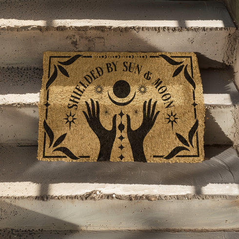 Natural coir doormat featuring black silhouettes of two open hands reaching toward a crescent moon and sun, surrounded by celestial and botanical details. The phrase Shielded by Sun & Moon is hand-stenciled above, with a non-slip vinyl backing for outdoor use.