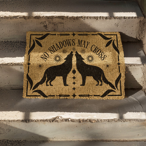 Natural coir doormat featuring black silhouettes of two howling wolves in symmetrical formation, surrounded by celestial and botanical details. The phrase No Shadows May Cross is hand-stenciled above, with a non-slip vinyl backing for outdoor use.