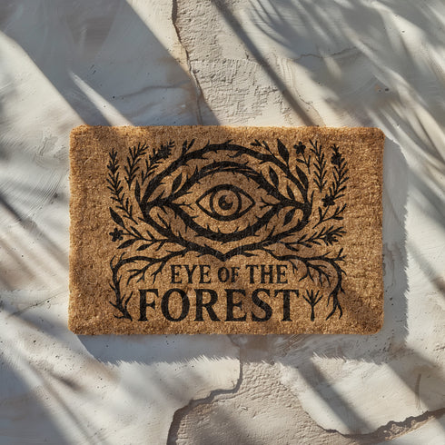 Rectangular coir doormat featuring black-ink artwork of a large all-seeing eye surrounded by branches, leaves, and forest flora. Beneath the design, the phrase “Eye of the Forest” is printed in bold serif font. Made from natural coconut coir with a vinyl backing, the mat measures 24 by 16 inches and is suitable for outdoor entryways.