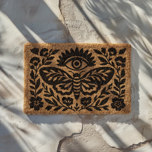A rectangular, natural brown coir doormat with a coarse, tufted texture. The design is printed in solid black ink in a folk-art, block-print style. In the center is a large, stylized death's-head moth with intricate patterns on its wings. Above the moth is a central all-seeing eye with radiating lashes. The entire central image is enclosed by a symmetrical botanical frame of stylized flowers, stems, and leaves, creating a decorative border. The doormat has a non-slip black vinyl backing.