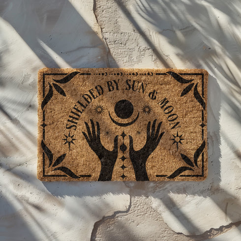 Natural coir doormat featuring black silhouettes of two open hands reaching toward a crescent moon and sun, surrounded by celestial and botanical details. The phrase Shielded by Sun & Moon is hand-stenciled above, with a non-slip vinyl backing for outdoor use.