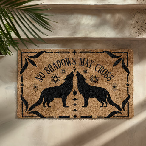 Natural coir doormat featuring black silhouettes of two howling wolves in symmetrical formation, surrounded by celestial and botanical details. The phrase No Shadows May Cross is hand-stenciled above, with a non-slip vinyl backing for outdoor use.