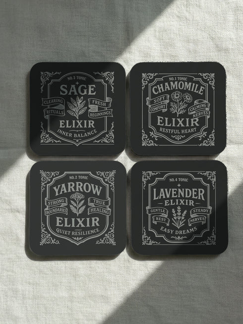 A complete set of four square, black, gloss beverage coasters with protective cork bases. Each coaster features an intricate Art Nouveau botanical design in white, stylized as a vintage elixir label. The set includes distinct labels for Sage, Chamomile, Yarrow, and Lavender, each inscribed with positive affirmations relating to healing and inner balance. These elegant Gothic drinkware pieces are designed to complement Dark Academia and Whimsigoth home decor, reflecting the arcane aesthetic.