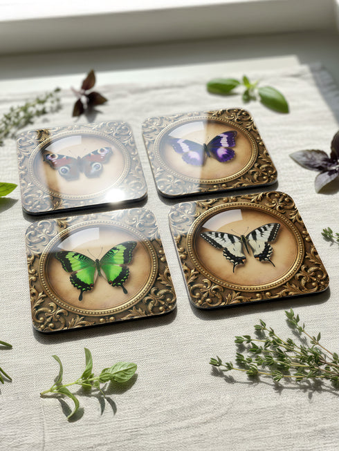 Grimoire and Goblet offers this luxury four-piece square Coaster Set featuring glossy, dome-covered, Taxidermy-inspired Entomology butterfly specimens, framed in ornate Victorian Gothic style. The resilient hardboard foundation and non-slip cork backing provide premium, waterproof protection for fine cocktails and esoteric teas. The collection includes purple, green, white, and peacock butterfly art, ideal for Dark Academia, Oddity, and Curio Cabinet decor.