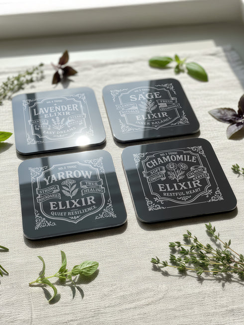 A complete set of four square, black, gloss beverage coasters with protective cork bases. Each coaster features an intricate Art Nouveau botanical design in white, stylized as a vintage elixir label. The set includes distinct labels for Sage, Chamomile, Yarrow, and Lavender, each inscribed with positive affirmations relating to healing and inner balance. These elegant Gothic drinkware pieces are designed to complement Dark Academia and Whimsigoth home decor, reflecting the arcane aesthetic.