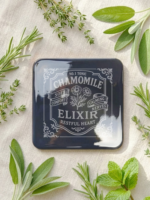 A complete set of four square, black, gloss beverage coasters with protective cork bases. Each coaster features an intricate Art Nouveau botanical design in white, stylized as a vintage elixir label. The set includes distinct labels for Sage, Chamomile, Yarrow, and Lavender, each inscribed with positive affirmations relating to healing and inner balance. These elegant Gothic drinkware pieces are designed to complement Dark Academia and Whimsigoth home decor, reflecting the arcane aesthetic.