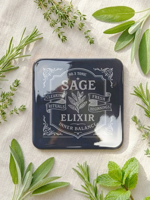 A complete set of four square, black, gloss beverage coasters with protective cork bases. Each coaster features an intricate Art Nouveau botanical design in white, stylized as a vintage elixir label. The set includes distinct labels for Sage, Chamomile, Yarrow, and Lavender, each inscribed with positive affirmations relating to healing and inner balance. These elegant Gothic drinkware pieces are designed to complement Dark Academia and Whimsigoth home decor, reflecting the arcane aesthetic.