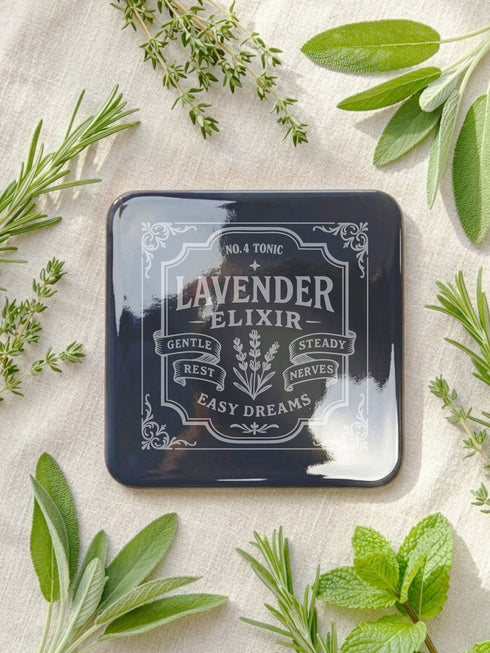 A complete set of four square, black, gloss beverage coasters with protective cork bases. Each coaster features an intricate Art Nouveau botanical design in white, stylized as a vintage elixir label. The set includes distinct labels for Sage, Chamomile, Yarrow, and Lavender, each inscribed with positive affirmations relating to healing and inner balance. These elegant Gothic drinkware pieces are designed to complement Dark Academia and Whimsigoth home decor, reflecting the arcane aesthetic.