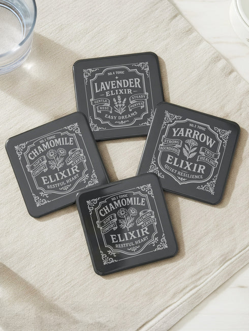 A complete set of four square, black, gloss beverage coasters with protective cork bases. Each coaster features an intricate Art Nouveau botanical design in white, stylized as a vintage elixir label. The set includes distinct labels for Sage, Chamomile, Yarrow, and Lavender, each inscribed with positive affirmations relating to healing and inner balance. These elegant Gothic drinkware pieces are designed to complement Dark Academia and Whimsigoth home decor, reflecting the arcane aesthetic.