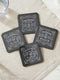 A complete set of four square, black, gloss beverage coasters with protective cork bases. Each coaster features an intricate Art Nouveau botanical design in white, stylized as a vintage elixir label. The set includes distinct labels for Sage, Chamomile, Yarrow, and Lavender, each inscribed with positive affirmations relating to healing and inner balance. These elegant Gothic drinkware pieces are designed to complement Dark Academia and Whimsigoth home decor, reflecting the arcane aesthetic.