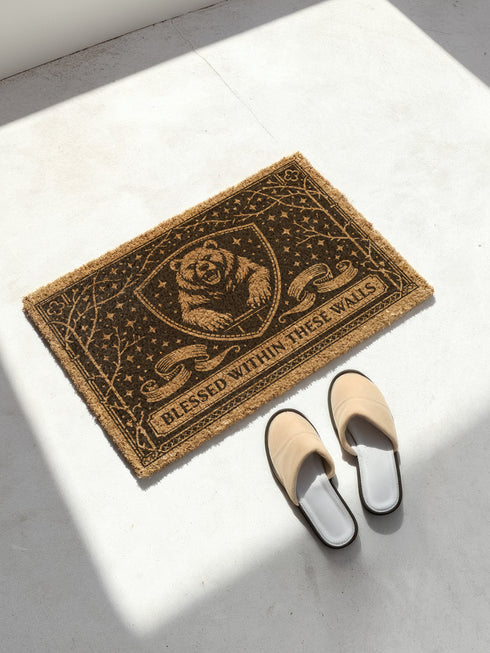 A rectangular eco-friendly coir doormat measuring 24 by 16 inches displayed on a stone porch. The natural golden-brown coconut fiber mat features a bold black graphic design of a heraldic bear head centered on a medieval knight's shield. Above and below the shield, Gothic-style typography reads BLESSED WITHIN THESE WALLS. The mat has a durable black vinyl non-slip backing. Ideal for Castlecore, Dark Academia, and Grimoire and Goblet style gothic home decor themes.