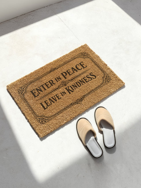 A high-angle close-up view of a Grimoire and Goblet luxury coir doormat. The mat features the phrase Enter in Peace Leave in Kindness painted in bold, Gothic-style black typography against the natural golden-brown texture of coconut husks. The text is enclosed in an intricate Victorian ornamental border with filigree corner flourishes. The texture appears coarse, durable, and dense, typical of high-quality tufted coconut fiber entry mats. The design evokes a Dark Academia and witchy aesthetic.