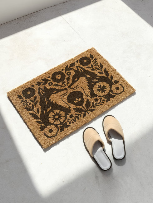 A rectangular, eco-friendly coir doormat with a natural, light brown tufted coconut fiber. Features a striking, symmetrical black folk-art design. In the center, two foxes face each other, noses touching, in a heraldic pose. They are surrounded by a dense, Scandinavian-style botanical border of stylized flowers, leaves, and branches, evoking a mystical woodland or dark cottagecore aesthetic. This witchy, Art Nouveau-inspired design serves as an elegant, gothic, or pagan piece of entryway decor.