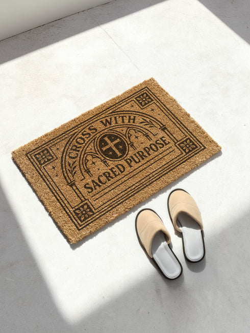 A rectangular eco-friendly coir doormat measuring 24×16 inches, shown on a dark stone entryway. Crafted from natural coconut fiber with a weather-resistant texture to grip dirt. Printed in bold black ink, it features a detailed Christian Gothic design: a central cross within an ornate circle, framed by cathedral arches. The top reads “CROSS WITH,” the bottom “SACRED PURPOSE,” all enclosed by a floral gothic border. Includes a black vinyl non-slip backing.