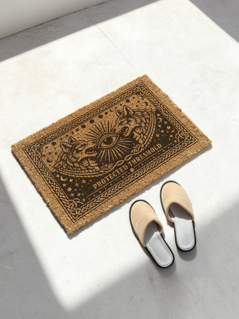 Grimoire and Goblet eco-friendly coir doormat featuring a mystical black wolf pack design. Two wolves face each other with a central all-seeing eye and celestial moon phases above. The text Protected Threshold anchors the bottom. A decorative celtic knot border frames the rug. The material is natural tufted coconut fiber with a non-slip black vinyl back. This gothic home decor piece serves as a spiritual ward and durable outdoor welcome mat for a witchy or dark academia entryway.