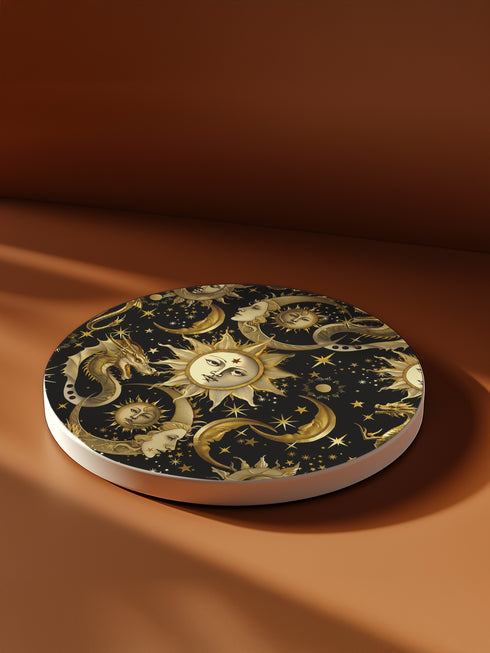 A round ceramic coaster featuring a dark navy background with golden celestial design elements: baroque-style sun faces, crescent moons with feminine profiles, stars, and a mystical dragon. The pattern is rendered in metallic gold tones, creating a rich, ornate astronomical motif.