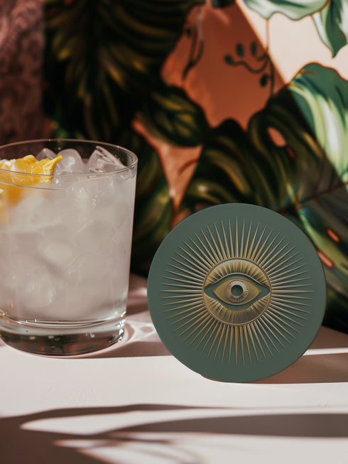 Round ceramic coaster with sage green background featuring a gold Evil Eye symbol in the center with radiating gold sunburst lines extending outward from the eye design.
