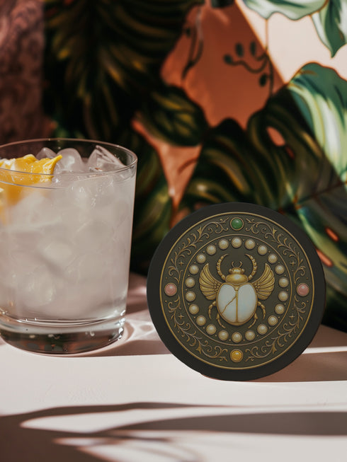Round ceramic coaster with a cork backing, featuring an ornate scarab beetle design surrounded by gold scrollwork, pearls, and gemstone accents in a circular motif.