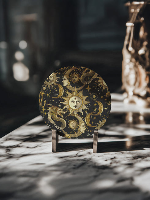 A round ceramic coaster featuring a dark navy background with golden celestial design elements: baroque-style sun faces, crescent moons with feminine profiles, stars, and a mystical dragon. The pattern is rendered in metallic gold tones, creating a rich, ornate astronomical motif.